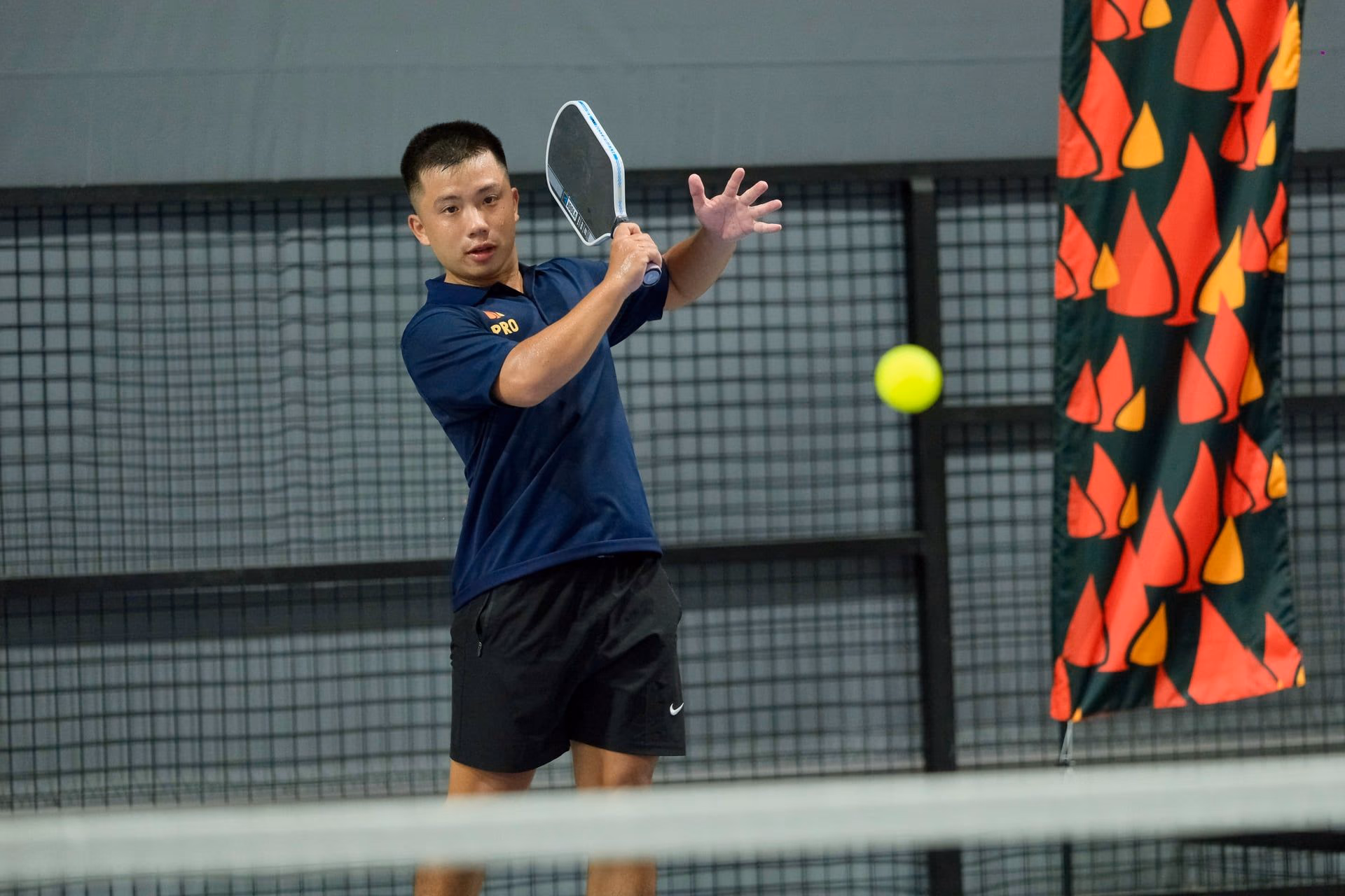 A coach in a Training Club polo mid-swing on a backhand drive, ball in flight.