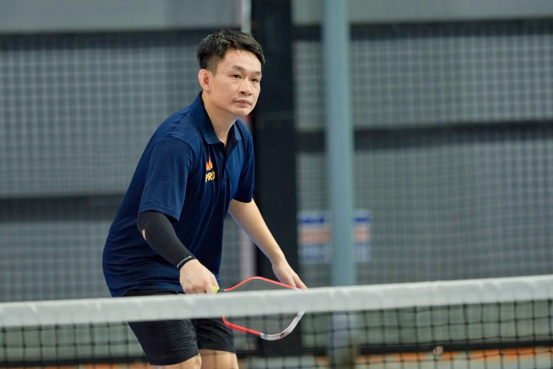 A coach in a Training Club polo at the net, paddle ready, watching the point.