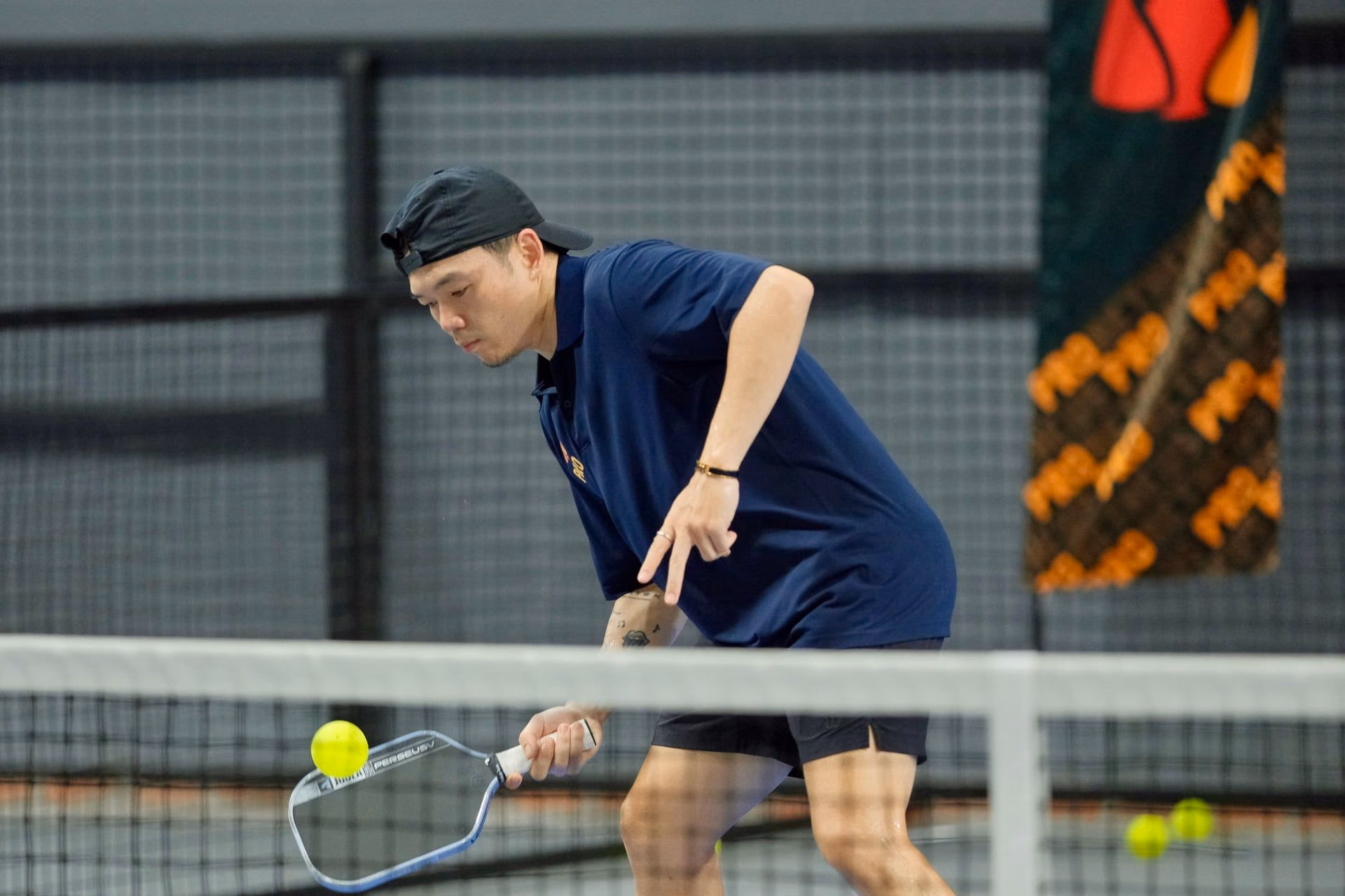 A coach in a Training Club polo bent low at the net, paddle out for a dink.