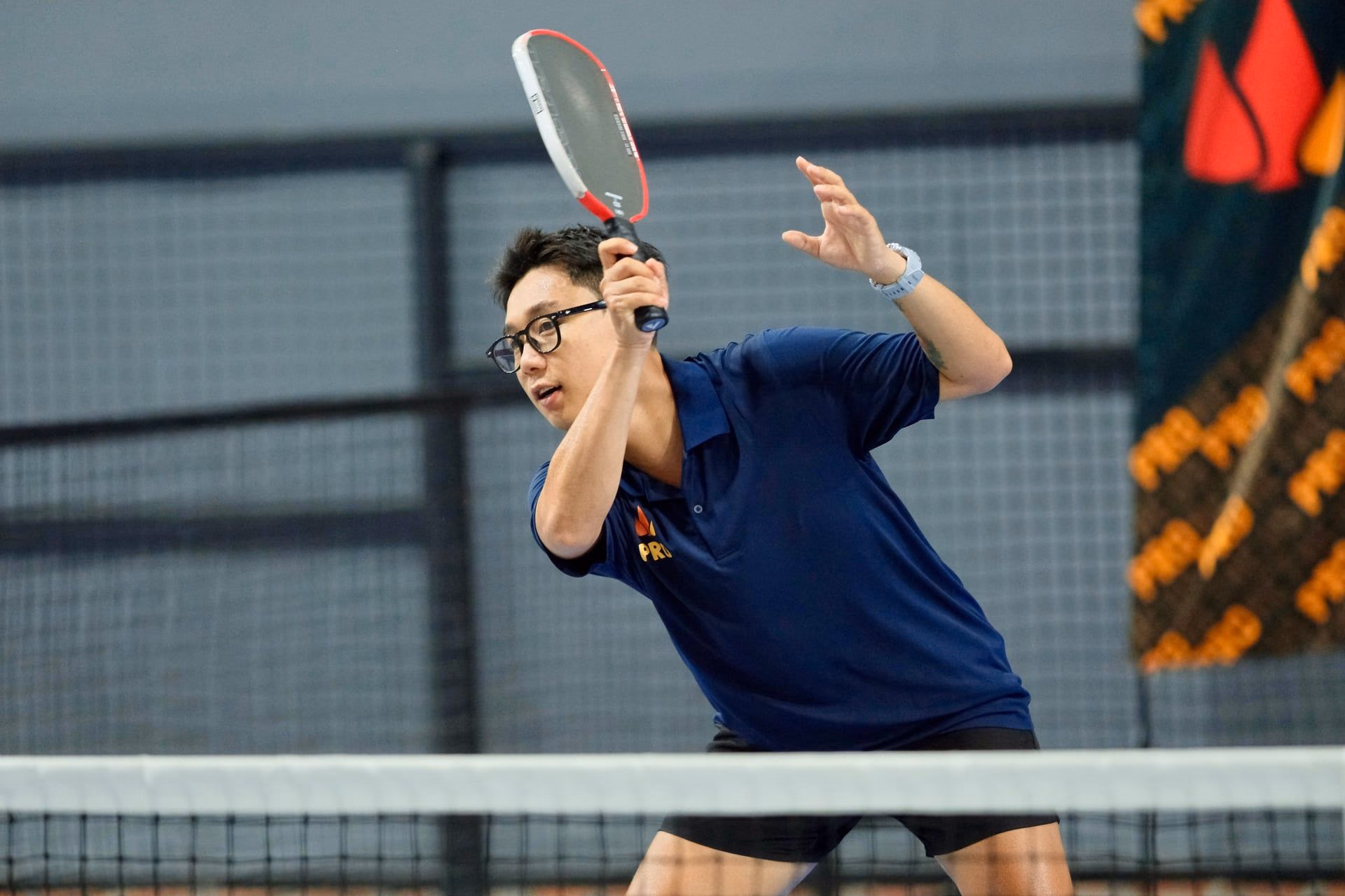 A coach in a Training Club polo, glasses on, mid-swing at the net with the ball in flight.